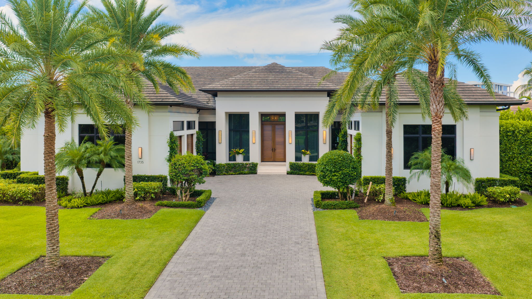 Modern single-story house with a gray shingle roof, large wooden front door, and tall windows, flanked by symmetrical green landscaping with palm trees and trimmed bushes along a wide paved driveway.
