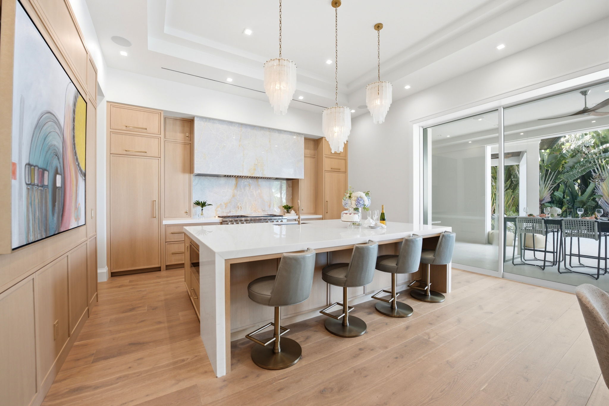 Modern kitchen with light wood cabinetry, large white marble island with four gray bar stools, three pendant lights, and a sliding glass door opening to an outdoor dining area with greenery.