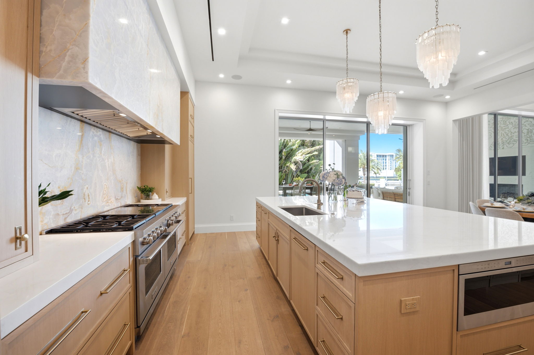 Modern kitchen with light wood cabinets, large white marble island with built-in sink, three hanging chandeliers, stainless steel oven and stovetop, and sliding glass doors opening to an outdoor patio with plants and seating.