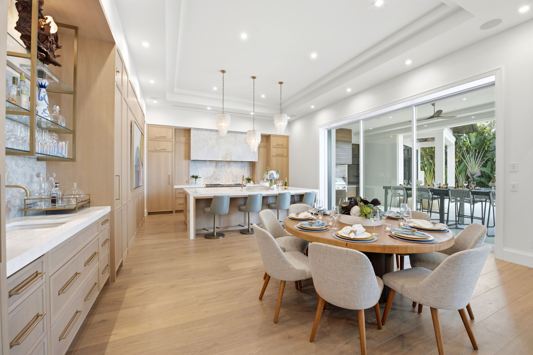 Modern dining area with a round wooden table set for six with plates and glasses, beige upholstered chairs, light wood flooring, an adjacent kitchen featuring light wood cabinetry, a white marble backsplash and countertop island with four gray bar stools, three pendant lights above, and large sliding glass doors opening to an outdoor dining space surrounded by greenery.