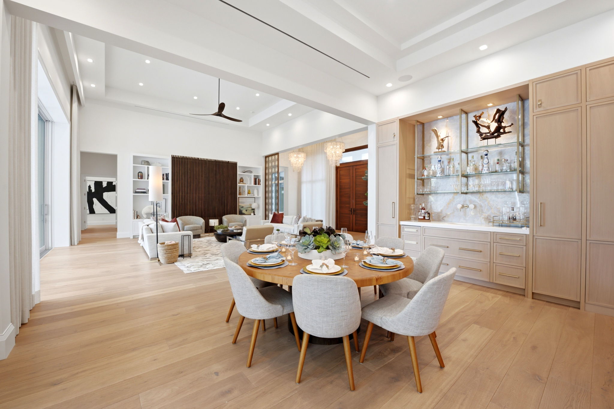 Bright open-concept dining and living area with light wood flooring, a round wooden dining table set for six with gray upholstered chairs, and a built-in bar with glass shelves and liquor bottles on the right. The living area in the background features neutral-toned sofas, armchairs, a rug, a floor lamp, and a decorative wooden accent wall. Large windows and double wooden doors provide natural light, and a modern ceiling fan hangs from the ceiling.