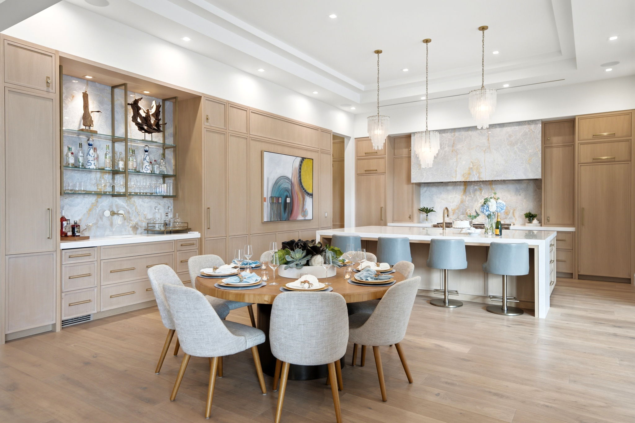 Bright modern kitchen and dining area with light wood cabinetry, a large white island with gold fixtures, three glass pendant lights, and four blue bar stools. A round wooden dining table with eight gray upholstered chairs is set with plates, napkins, and glassware. Decorative plants and art are displayed, with wood flooring throughout.