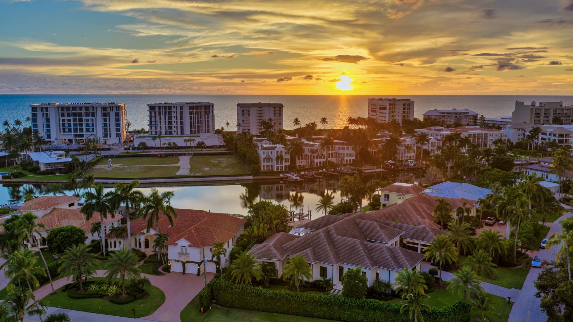 Aerial view of a coastal neighborhood at sunset featuring large houses with tiled roofs, palm trees, waterways, high-rise buildings near the ocean, and a vibrant sky with the sun setting over the water.