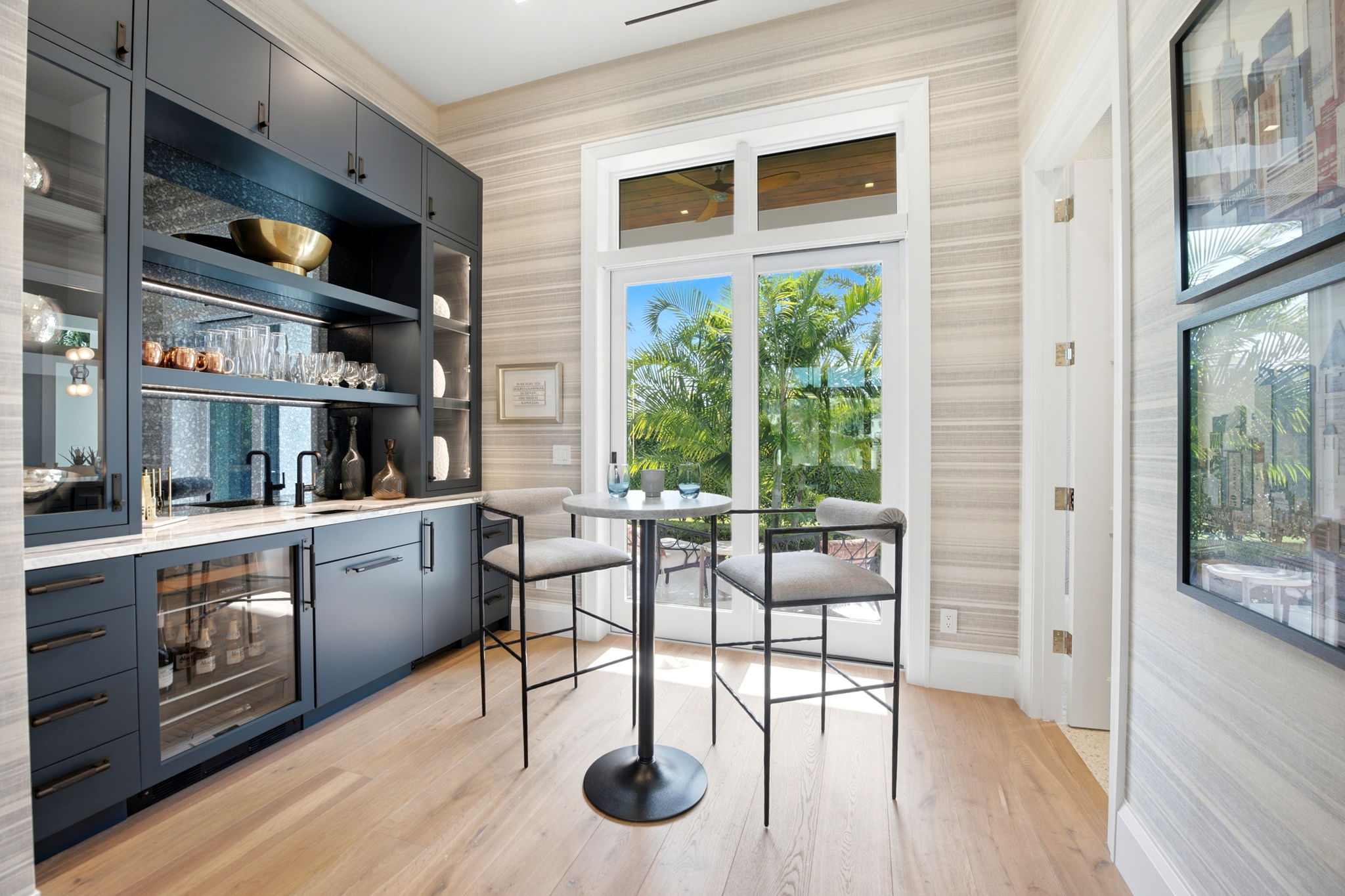 Modern dining nook with tall black metal chairs and round table set near glass doors revealing lush green palm trees outside, adjacent to navy blue cabinetry with glass shelves holding glasses and decorative items.