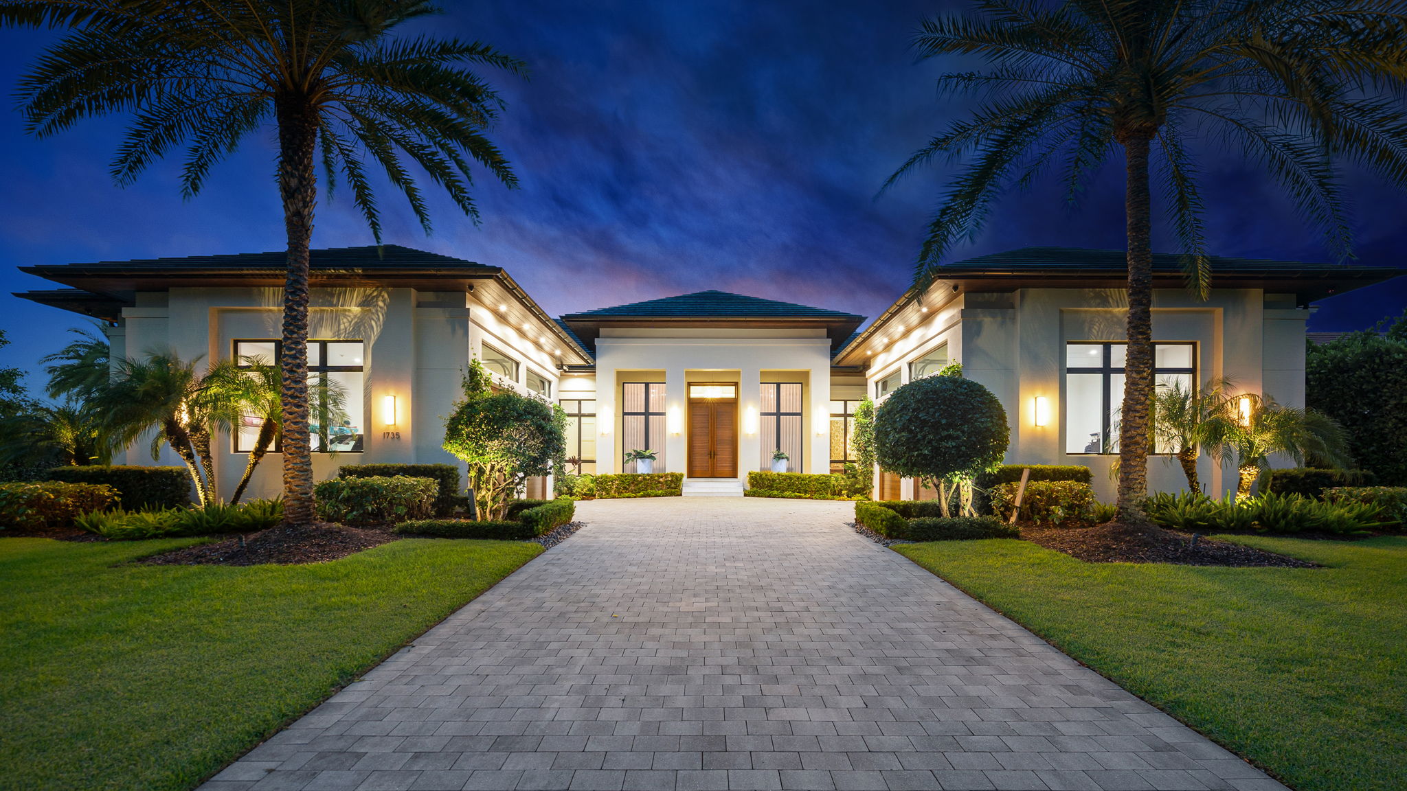 Night view of a modern house with a wide paved driveway leading to a wooden front door, flanked by large windows and trimmed hedges, palm trees, and exterior wall lights illuminating the facade and landscaping.