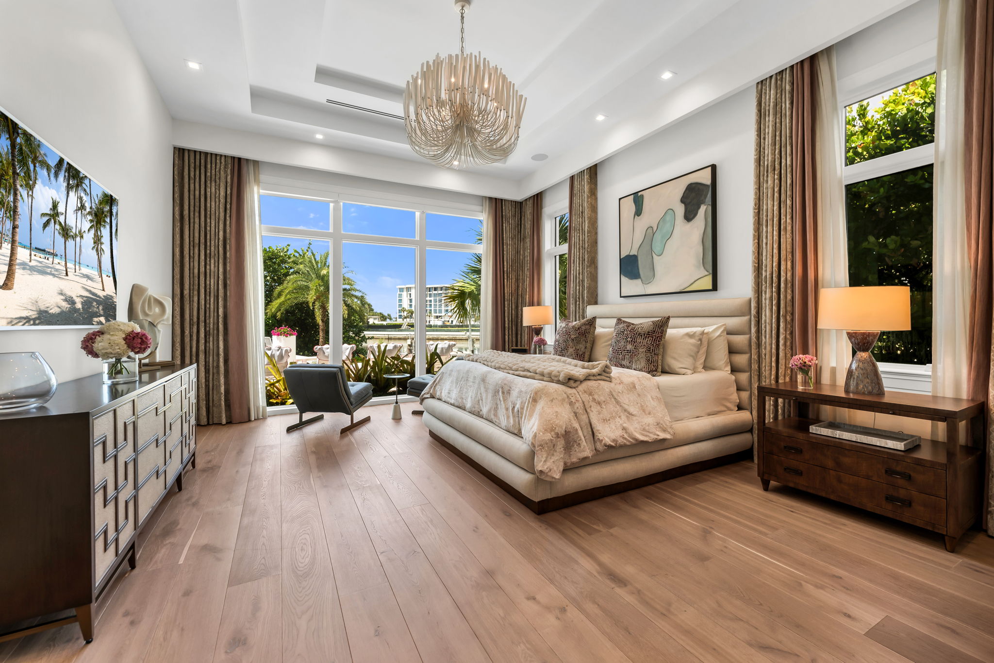 Modern bedroom with large windows showing greenery outside, featuring a padded bed with patterned pillows, wood flooring, a decorative chandelier, abstract wall art, dark wood furniture with lamps and floral arrangements, and a large flat-screen TV displaying a beach scene.