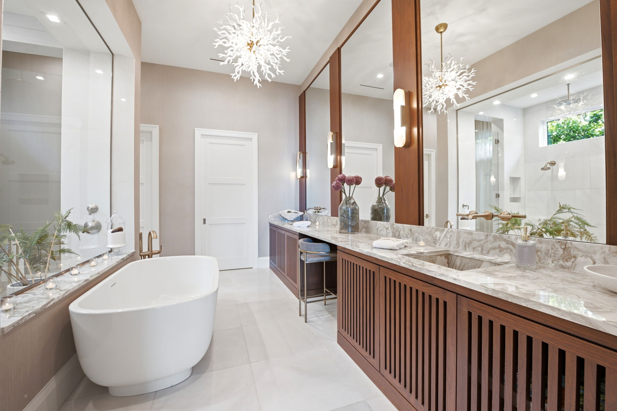 Modern bathroom with a freestanding white bathtub next to a large window, a long marble countertop with twin sinks, wooden cabinets, a cushioned stool, and decorative flowers in vases under large mirrors, lit by elegant white chandeliers.