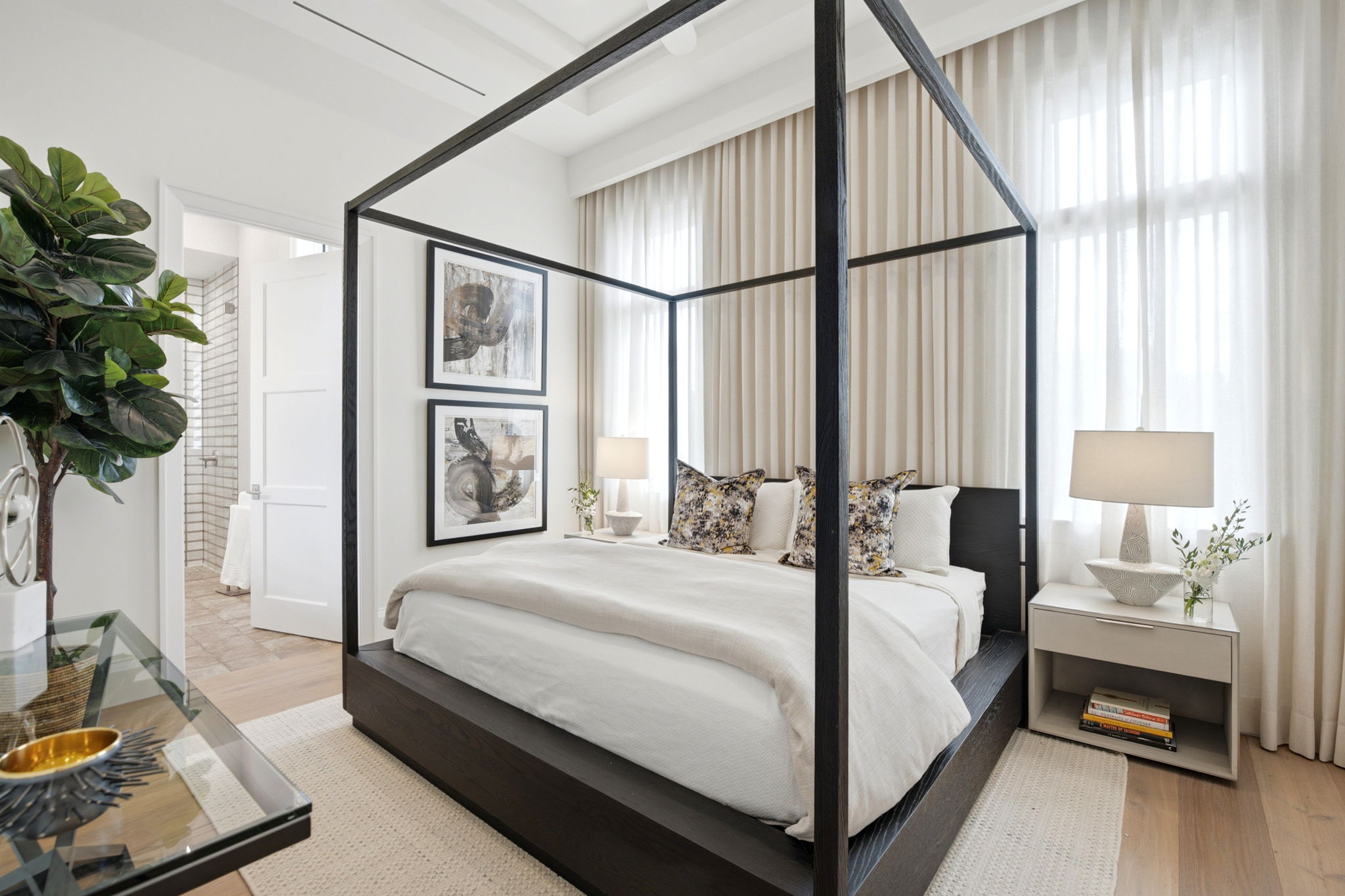 Modern bedroom with a black four-poster bed, white bedding, floral accent pillows, beige floor-to-ceiling curtains, two abstract paintings on the wall, nightstands with lamps, a plant, and an open door leading to a bathroom with tiled walls.
