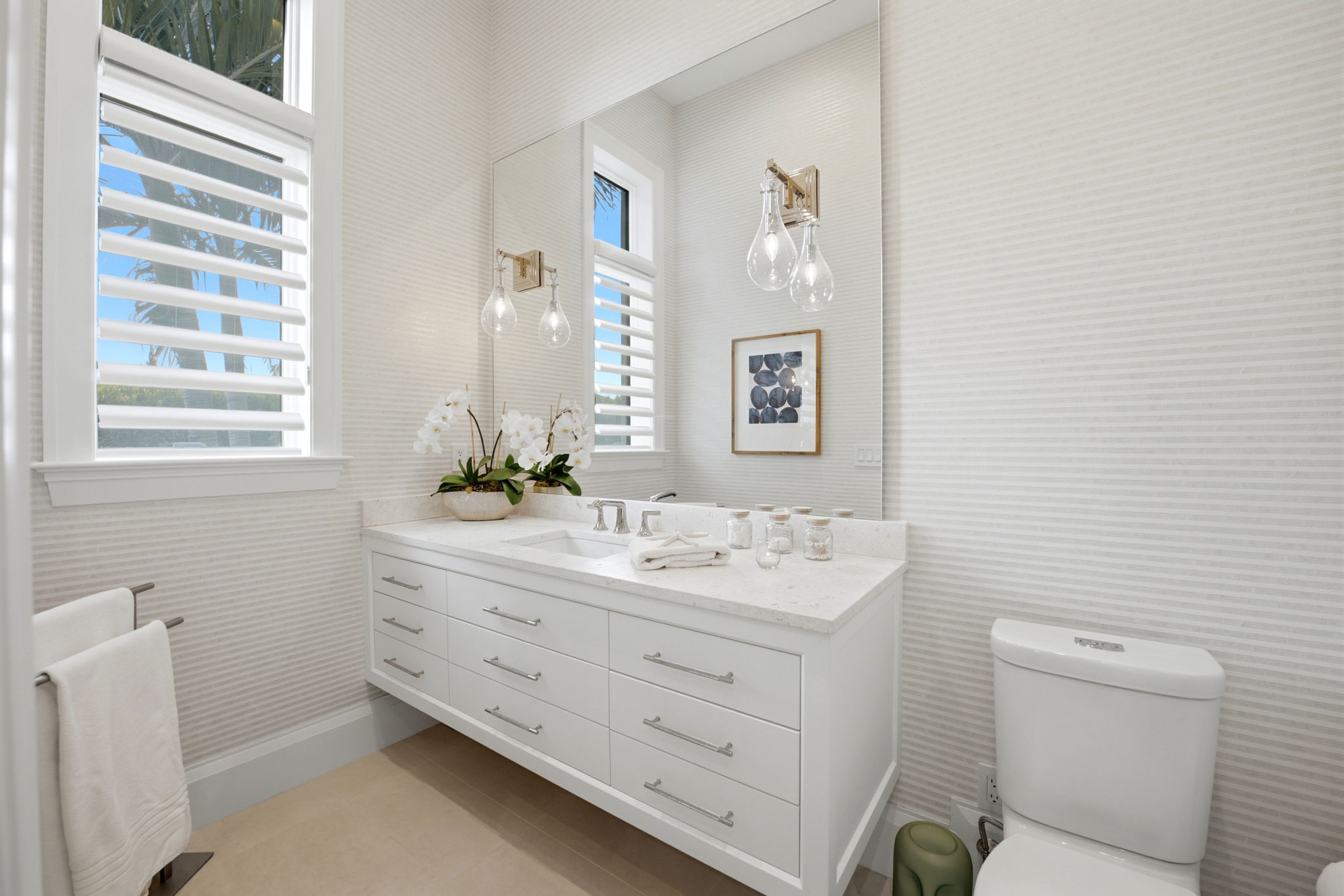 Bright modern bathroom with white floating vanity featuring multiple drawers, a large mirror, clear glass light fixtures, and a white orchid plant, next to a window with white shutters and a white toilet.