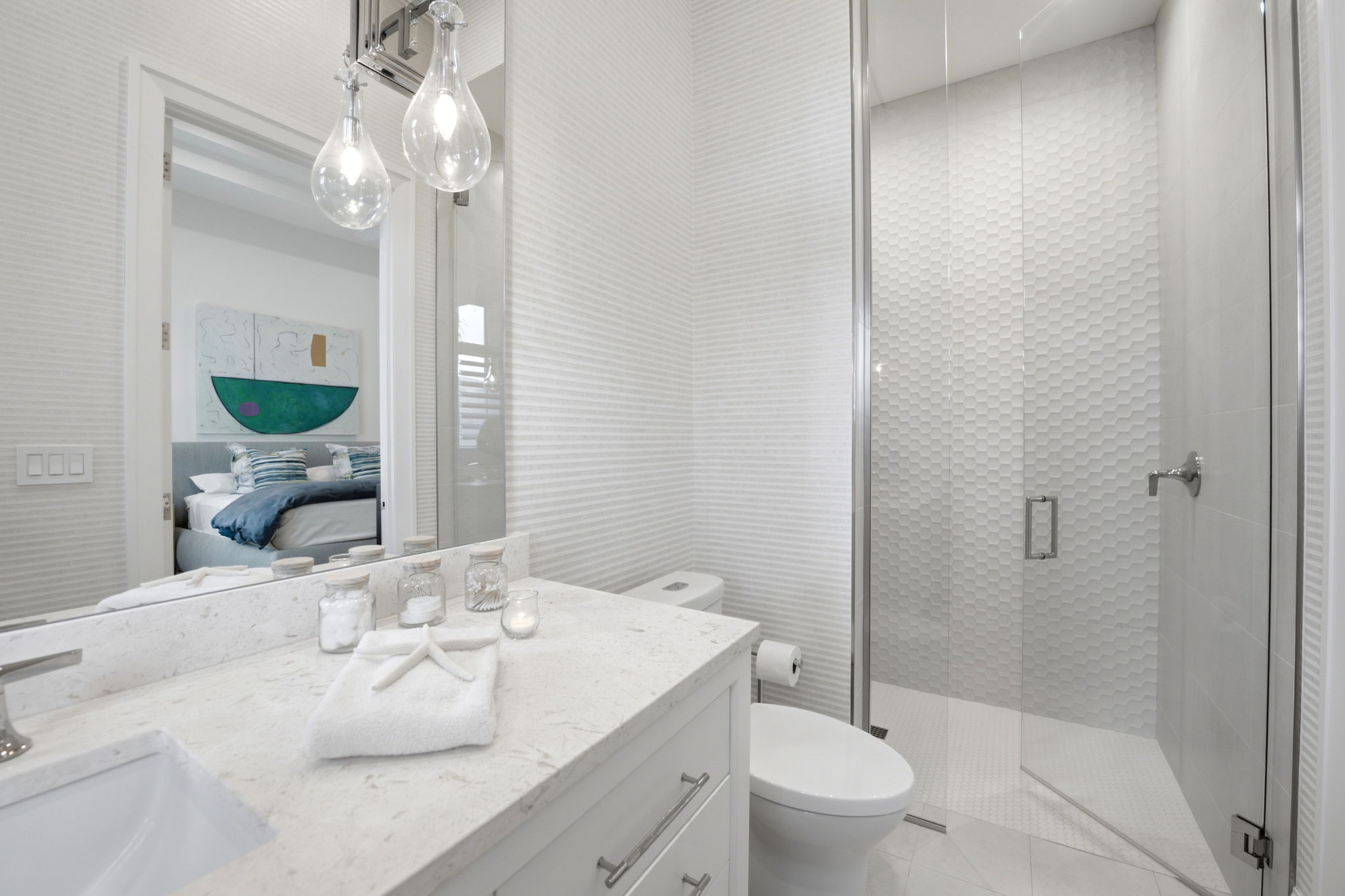 Modern bathroom with a white marble countertop, a folded towel with a decorative starfish, glass jars, a lit candle, a white toilet, and a glass-enclosed shower with textured hexagonal tile walls, reflecting a bedroom with a bed and abstract wall art.