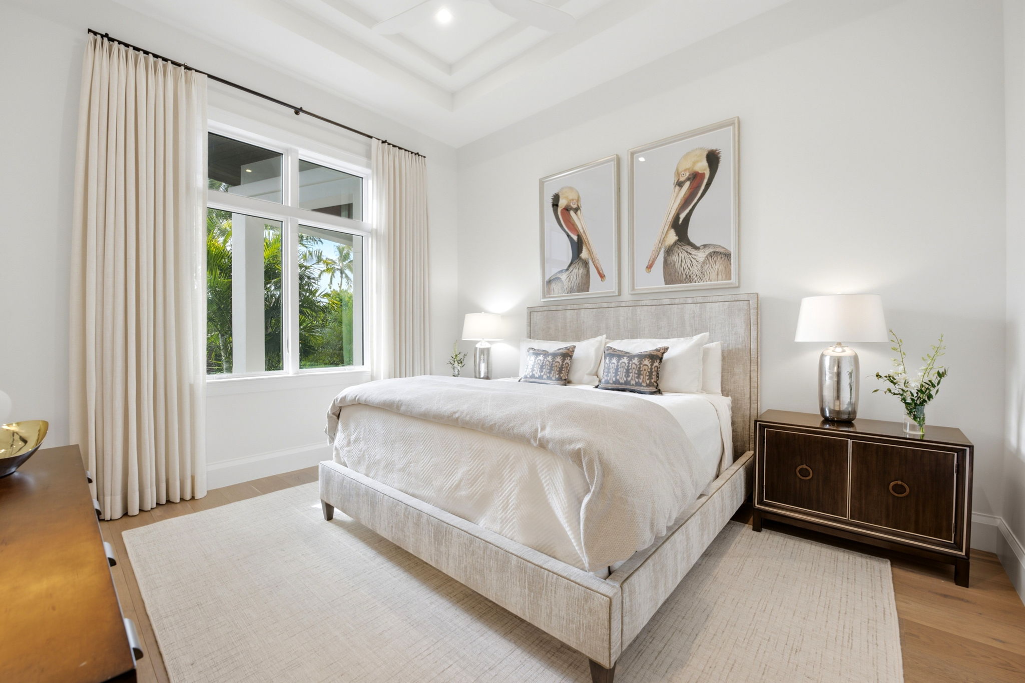 Bright bedroom with a large beige upholstered bed, two pillows with decorative covers, and two pelican artwork prints above the headboard, flanked by nightstands with silver lamps and small floral arrangements, next to a large window with cream curtains showing green palm trees outside.