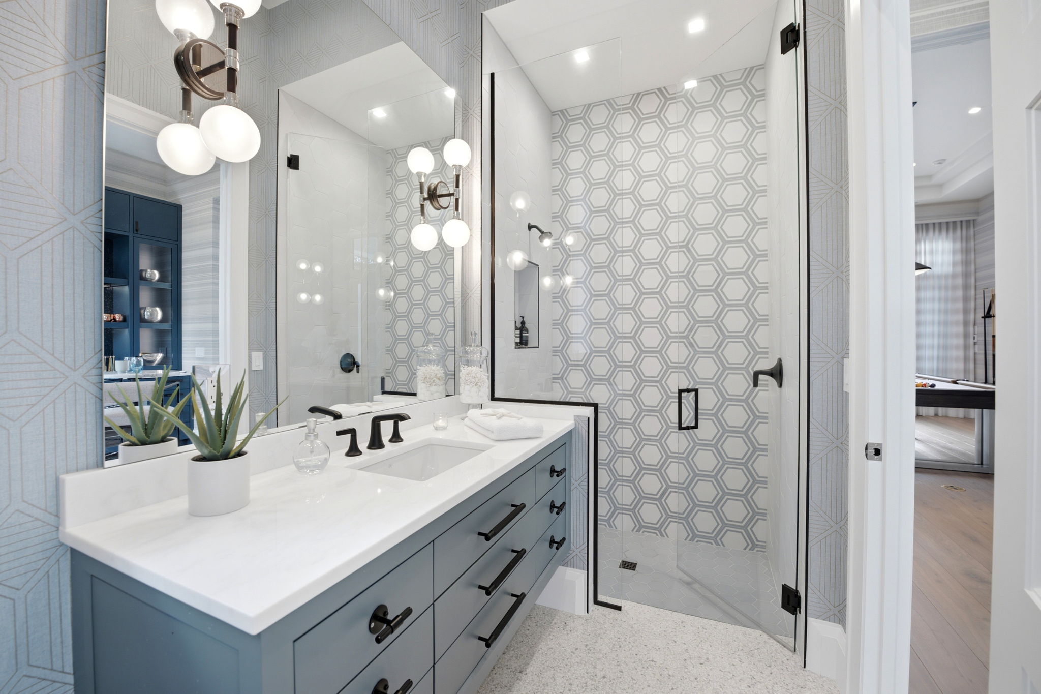 Modern bathroom with light blue vanity featuring black hardware, white countertop with sink, glass soap dispenser, and potted succulent, large mirror reflecting hexagonal patterned shower tiles with black-framed clear glass shower door and matte black fixtures.