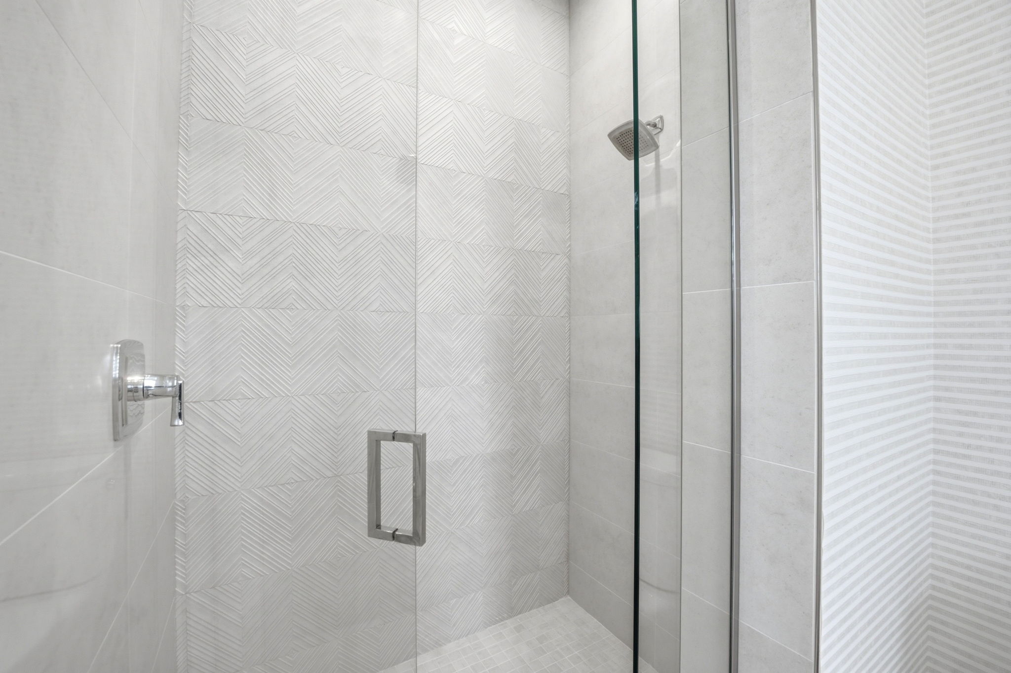 Minimalist walk-in shower with textured gray geometric wall tiles, clear glass door with metal handle, and a square overhead showerhead.