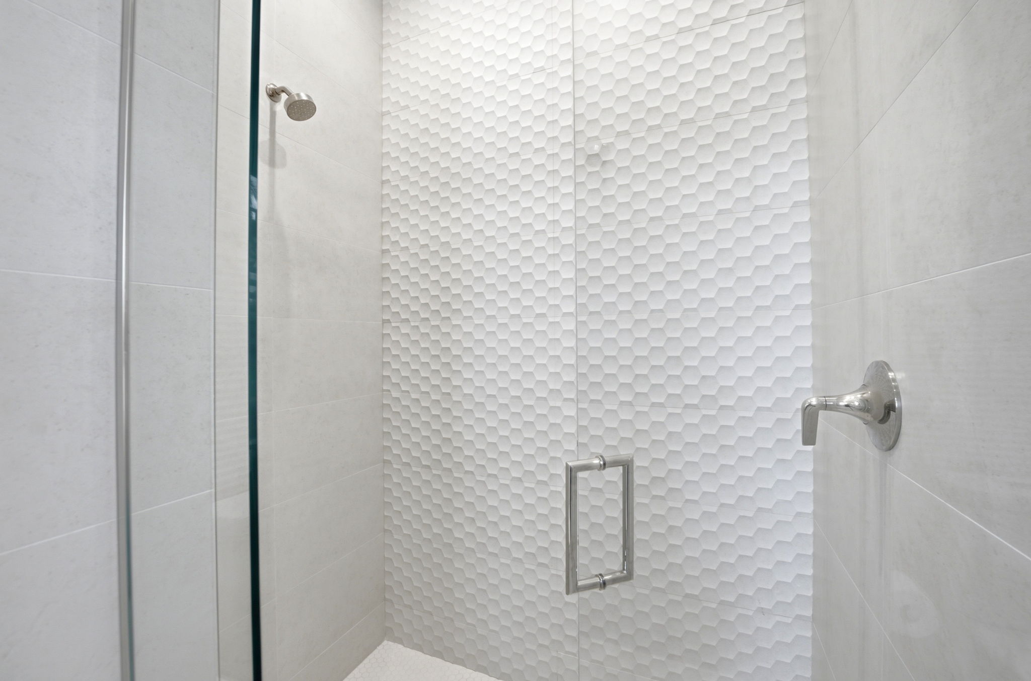 Minimalist walk-in shower with light gray large rectangular tiles, a glass door with silver handle, and a white textured hexagonal tile accent wall behind the showerhead.