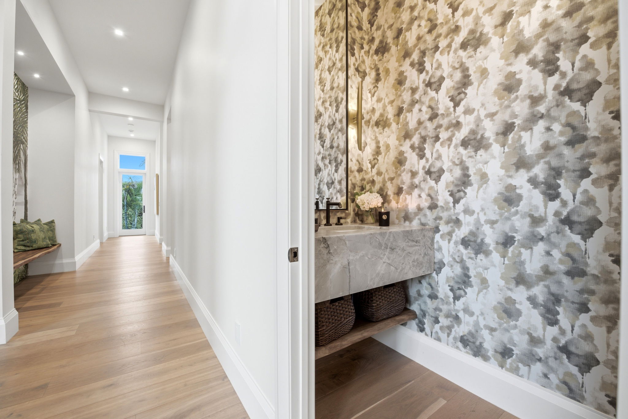 Modern hallway with light wood flooring and white walls, featuring a built-in bench with green pillows and a doorway leading to a powder room with patterned wallpaper, marble sink, mirror, and baskets underneath.