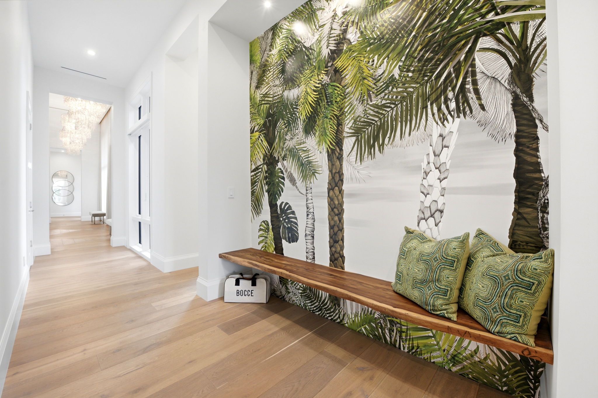 Bright hallway with light wood flooring, a long wooden bench mounted on the wall with three green patterned pillows, and a large mural of palm trees behind the bench.