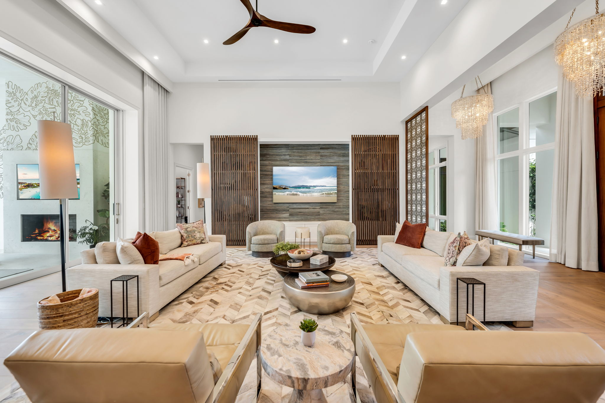 Bright, spacious modern living room with two beige sofas, two armchairs, a patterned rug, and a wall-mounted TV showing a beach scene, featuring wood accents, large windows, glass sliding doors, and a ceiling fan.
