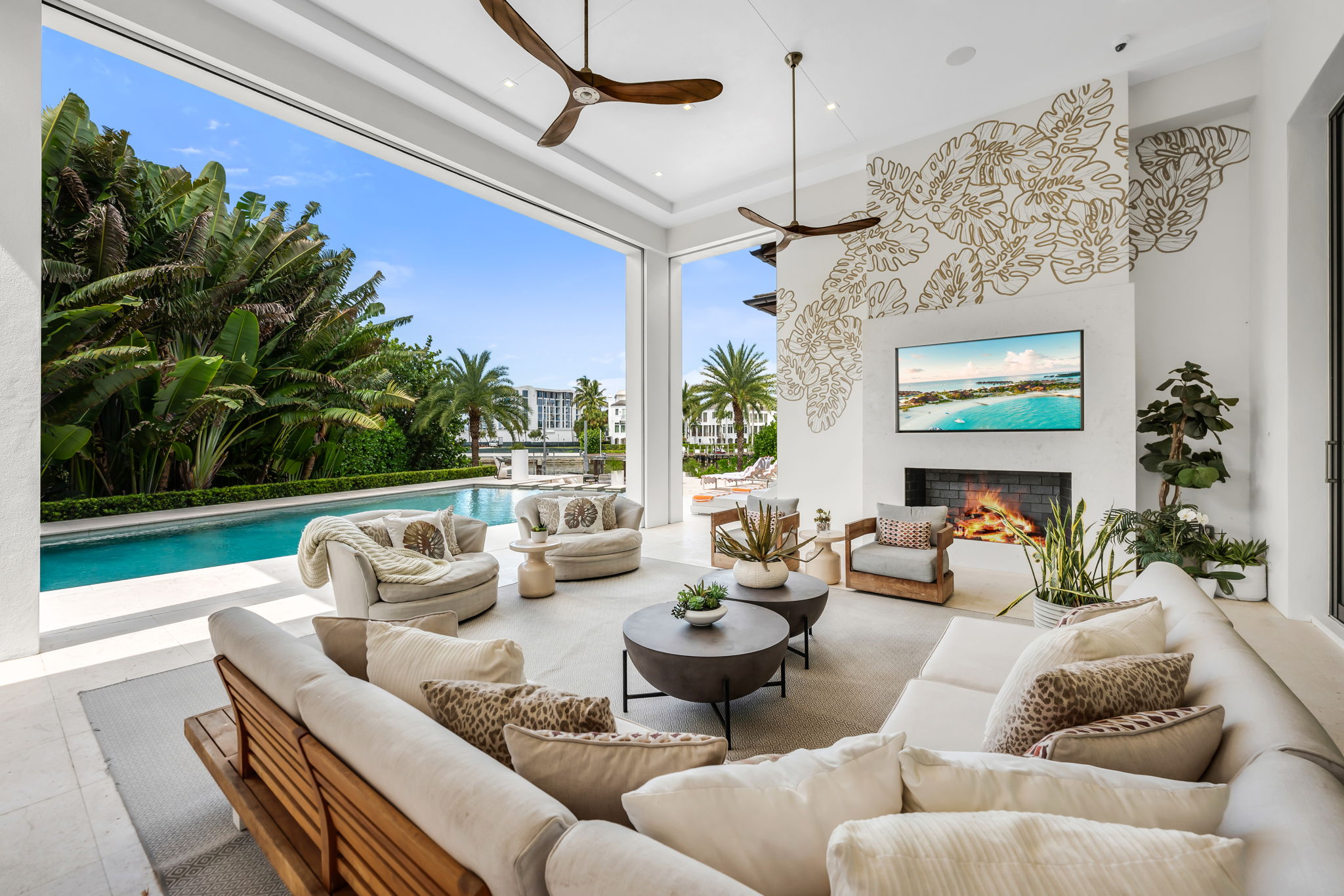 Modern outdoor living area with cream sectional sofa, two round coffee tables with plants, two circular armchairs, and a single armchair facing a built-in fireplace and wall-mounted TV, next to a swimming pool surrounded by tropical greenery and palm trees under a blue sky.