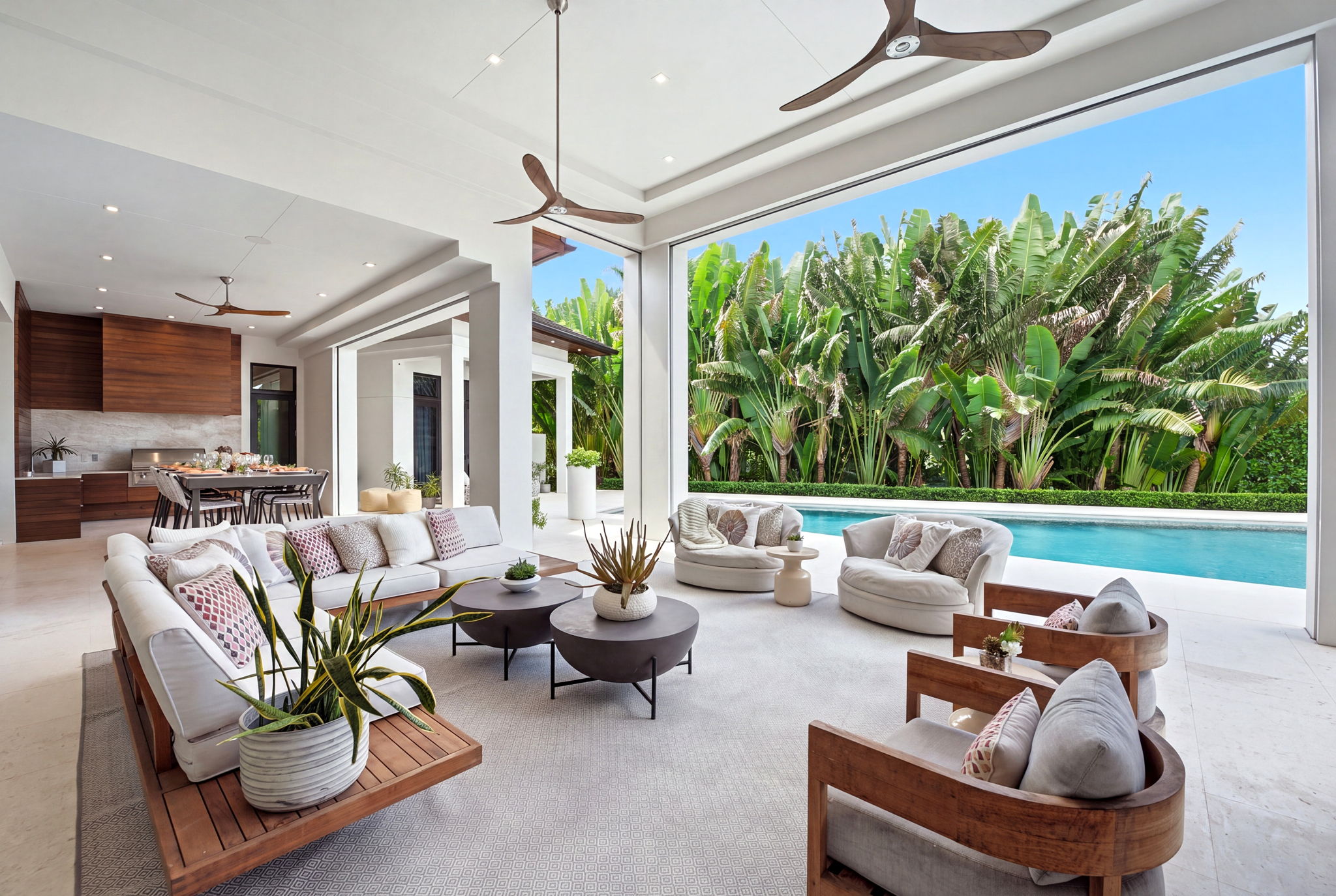 Open modern living area with wooden ceiling fans, white cushioned sofa and armchairs, round coffee tables with plants, dining table set for meal, and large open wall leading to a swimming pool and lush green tropical plants.