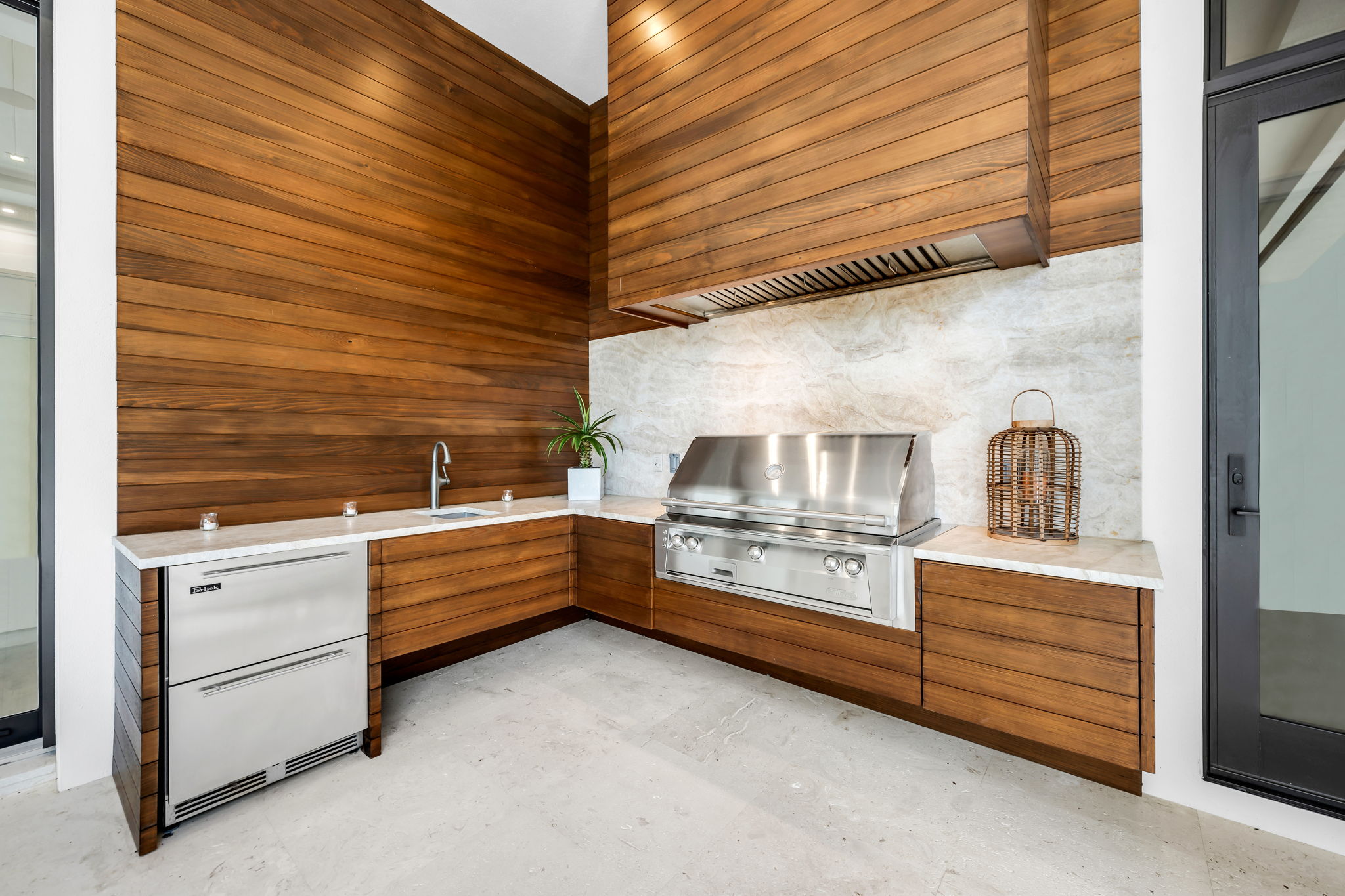Modern outdoor kitchen with wood paneling, white marble countertops, stainless steel built-in grill and refrigerator, small sink, potted plant, and decorative lantern on countertop near black-framed glass door.