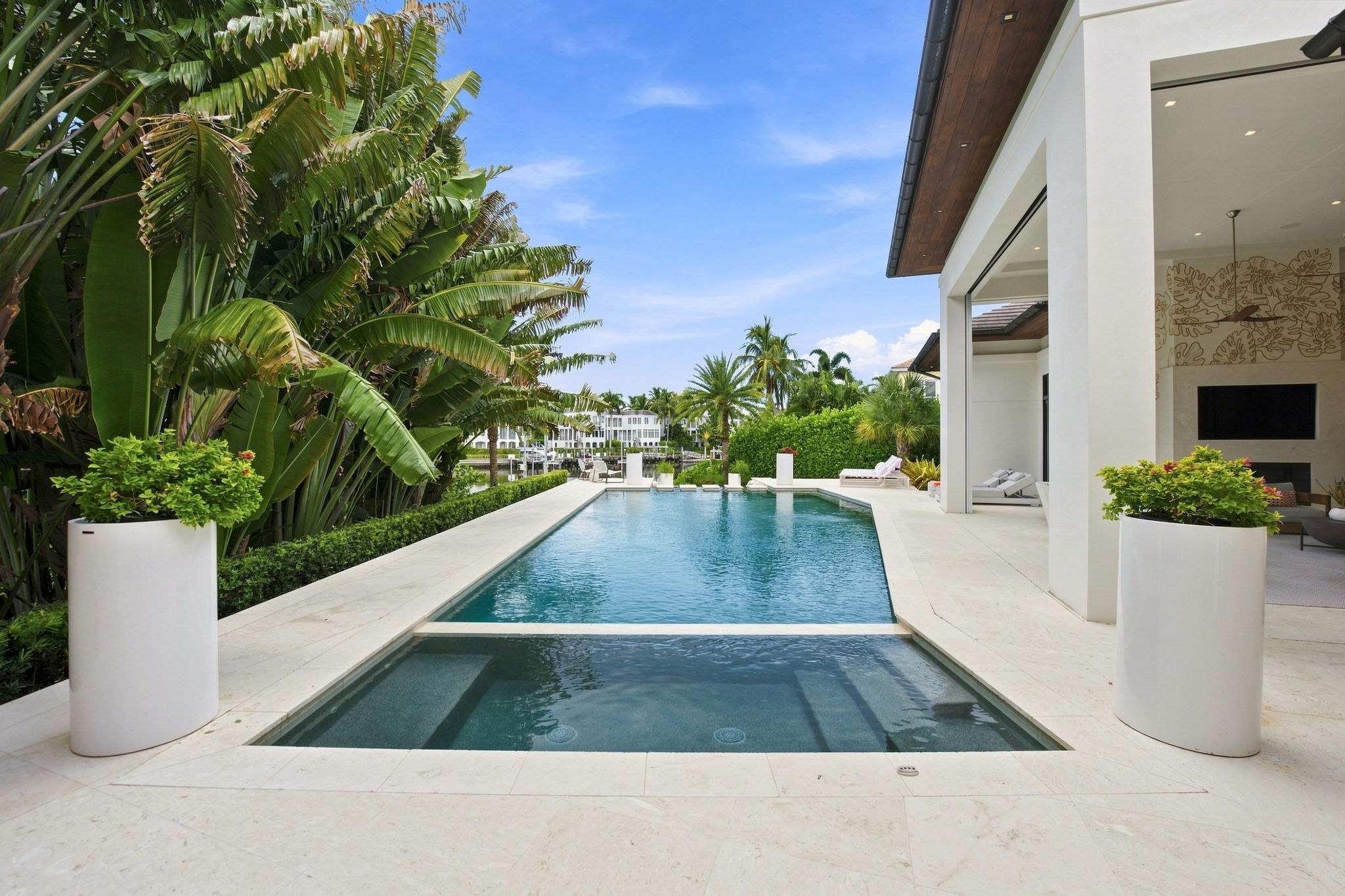 Modern outdoor pool with adjacent raised spa surrounded by light stone tiles, lush tropical plants on one side, and a white luxury house with an open lounge area on the other, under a clear blue sky.