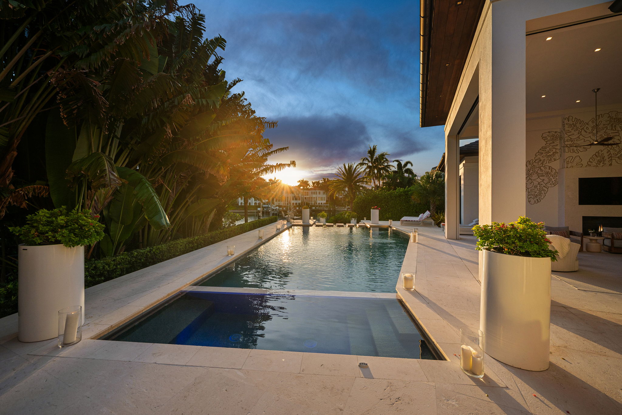Modern backyard pool at sunset with clear water, surrounded by large tropical plants and white stone decking, next to a stylish outdoor living area with open walls and seating.