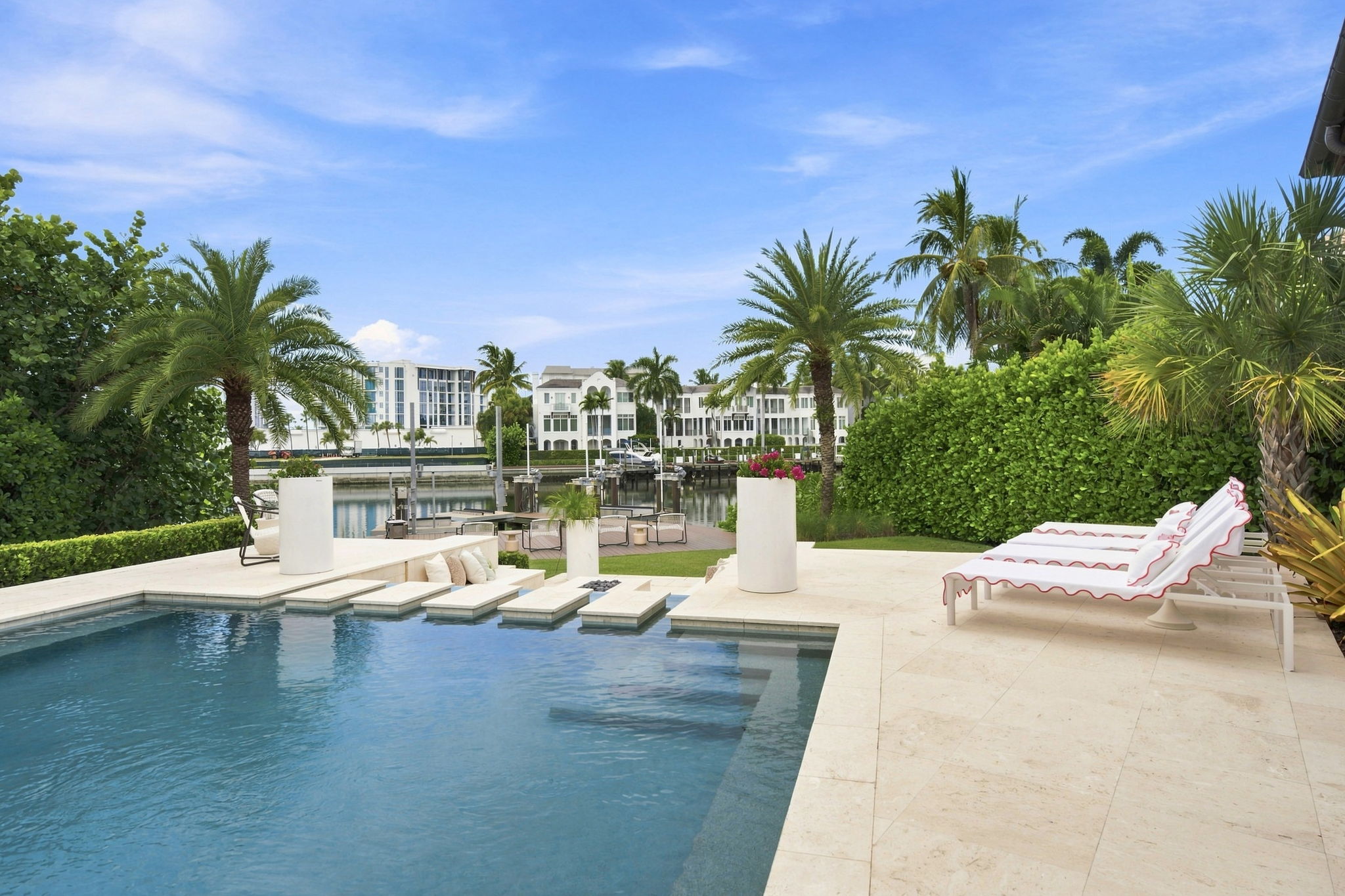 Luxurious outdoor pool area with beige stone deck, red-trimmed loungers, sunken seating, palm trees, and waterfront view of white buildings.