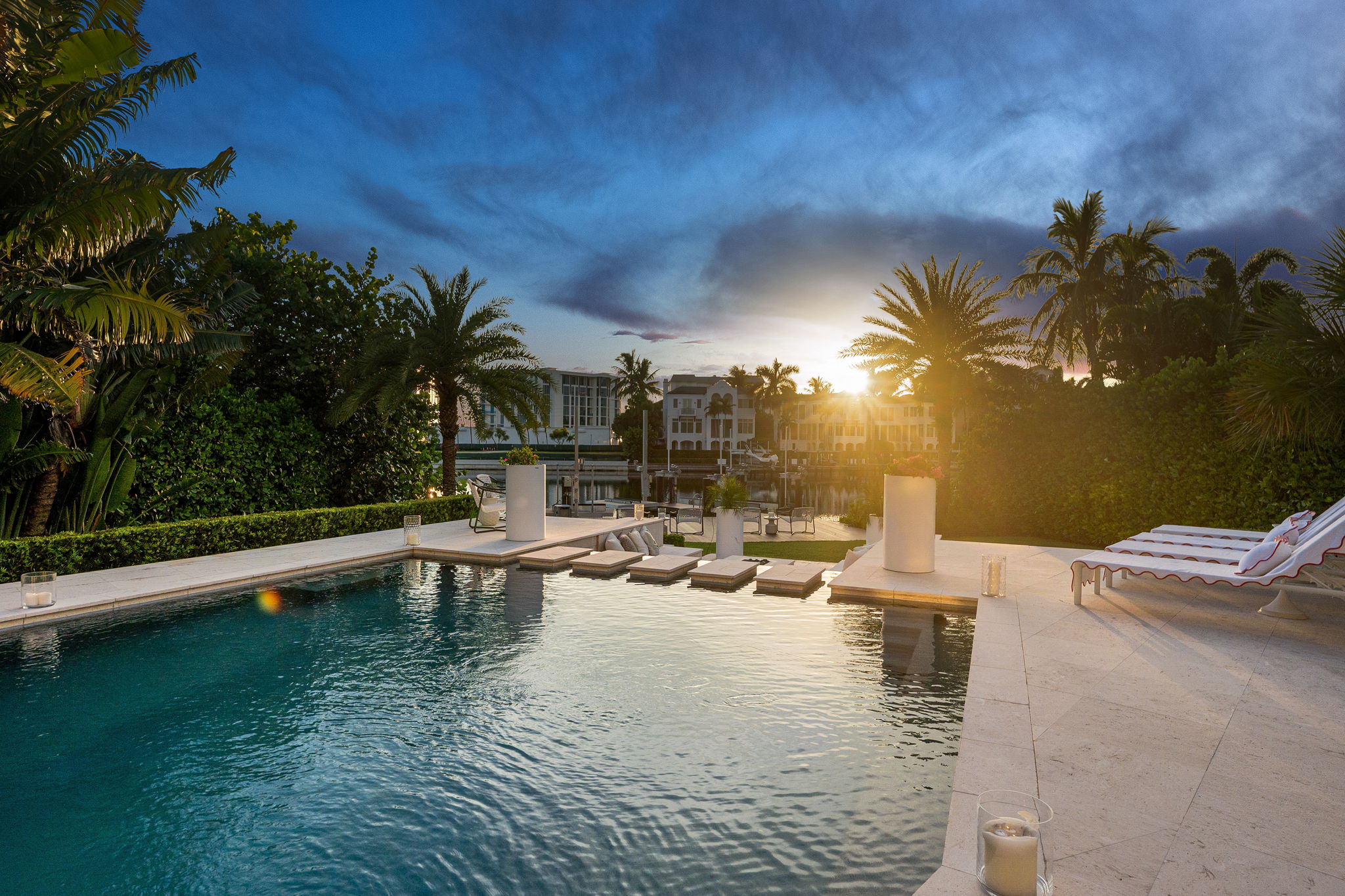Luxury outdoor pool area at sunset featuring stepping stones across the water, lounge chairs with white cushions and red trim, tall white planters with flowers, surrounded by palm trees and tropical plants, with calm water reflecting the sky and nearby waterfront homes.