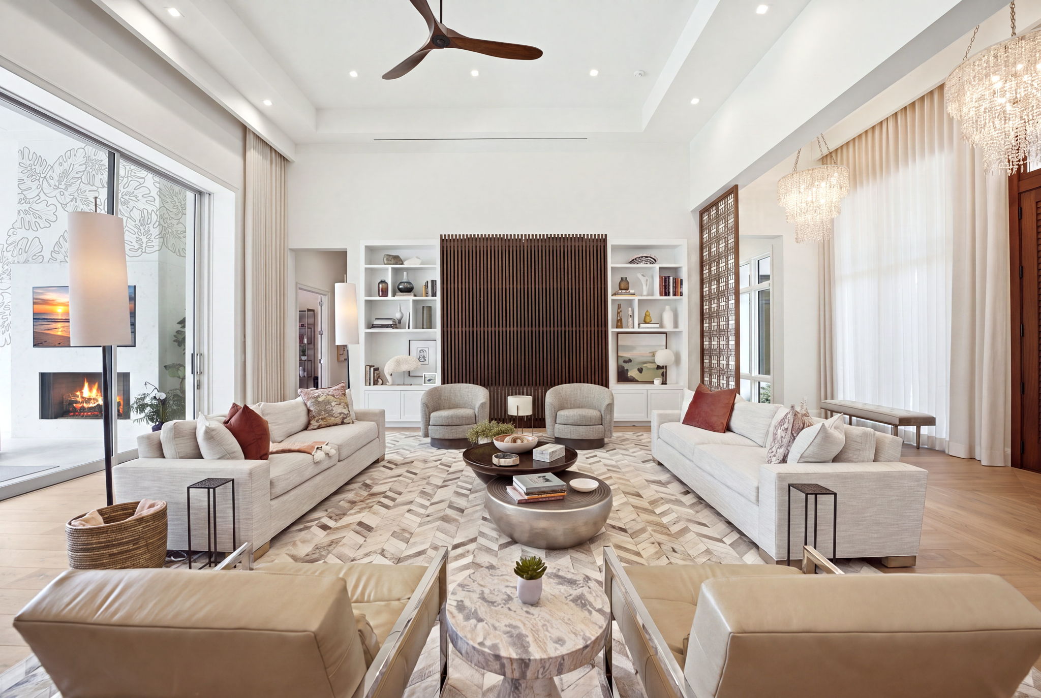 Spacious modern living room with high ceilings, two beige sofas facing each other, two beige armchairs, round coffee tables, built-in white shelves with décor and books, wooden slatted panel on back wall, floor-to-ceiling windows with curtains, two crystal chandeliers, and a ceiling fan. A basket, tall lamps, and a fireplace with a TV showing a sunset are visible on the left.