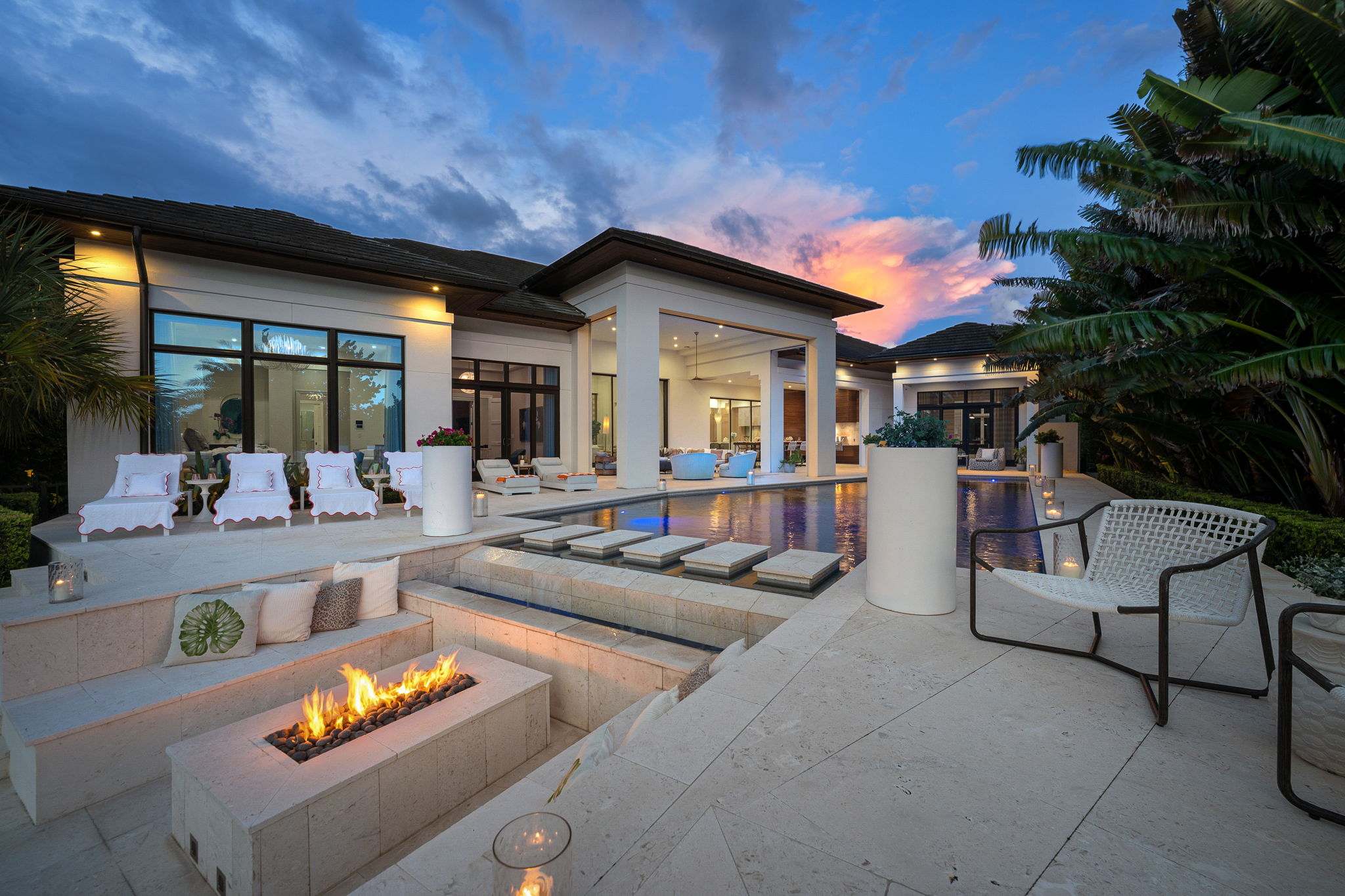 Modern luxury house exterior with a pool at sunset, featuring a sunken seating area with a fire pit, multiple white lounge chairs, large windows, and tropical plants.