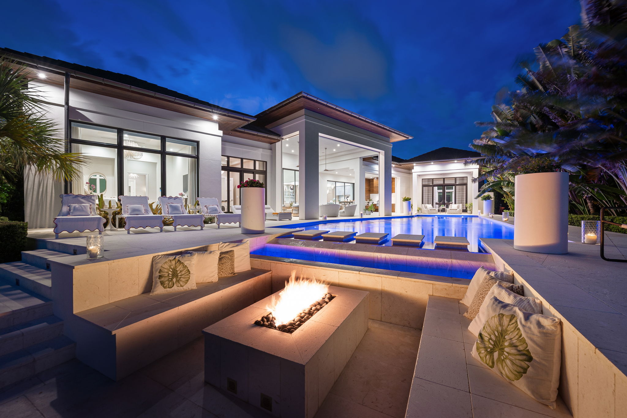 Modern luxurious house at night with a lit swimming pool featuring floating stepping stones, surrounded by white lounge chairs and a built-in sunken seating area with cushions around a rectangular fire pit, illuminated under a deep blue sky.