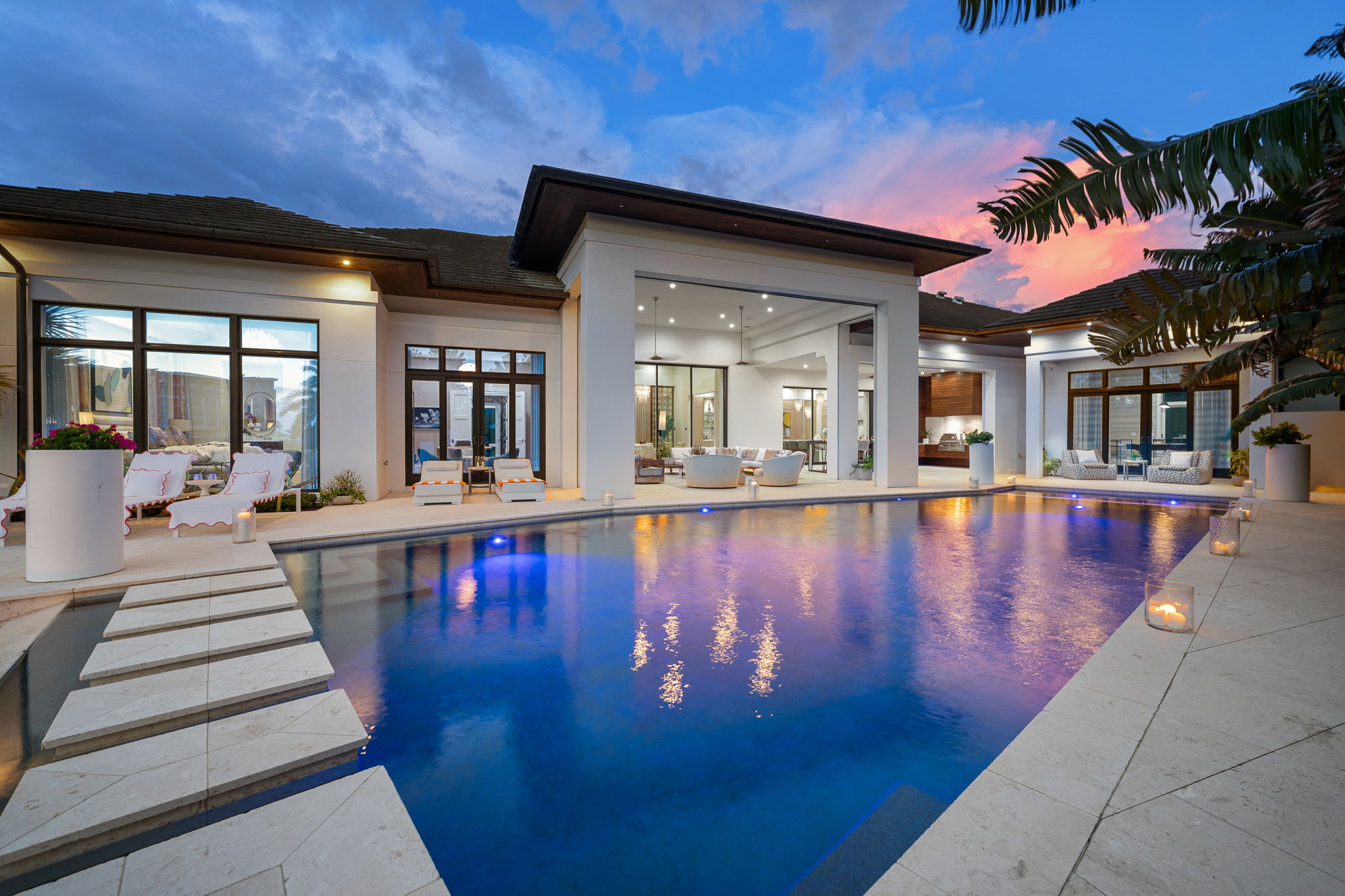 Luxurious modern home exterior at dusk with a large illuminated swimming pool reflecting pink and blue sky, white stone patio, reclining lounge chairs, outdoor seating areas, and tall plants.