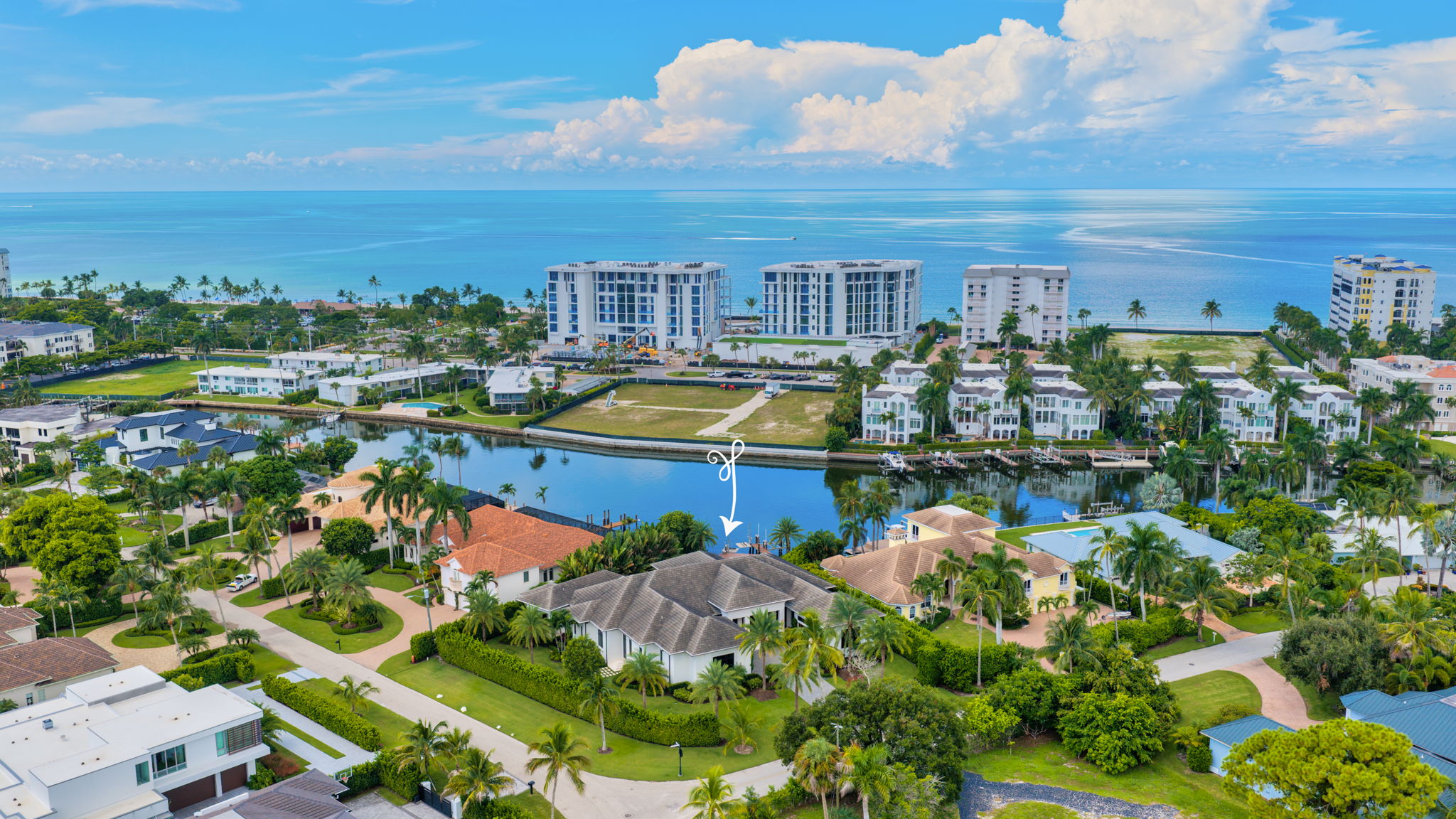 Aerial view of a coastal residential neighborhood with large houses, palm trees, a canal, and the ocean in the background under a partly cloudy blue sky.