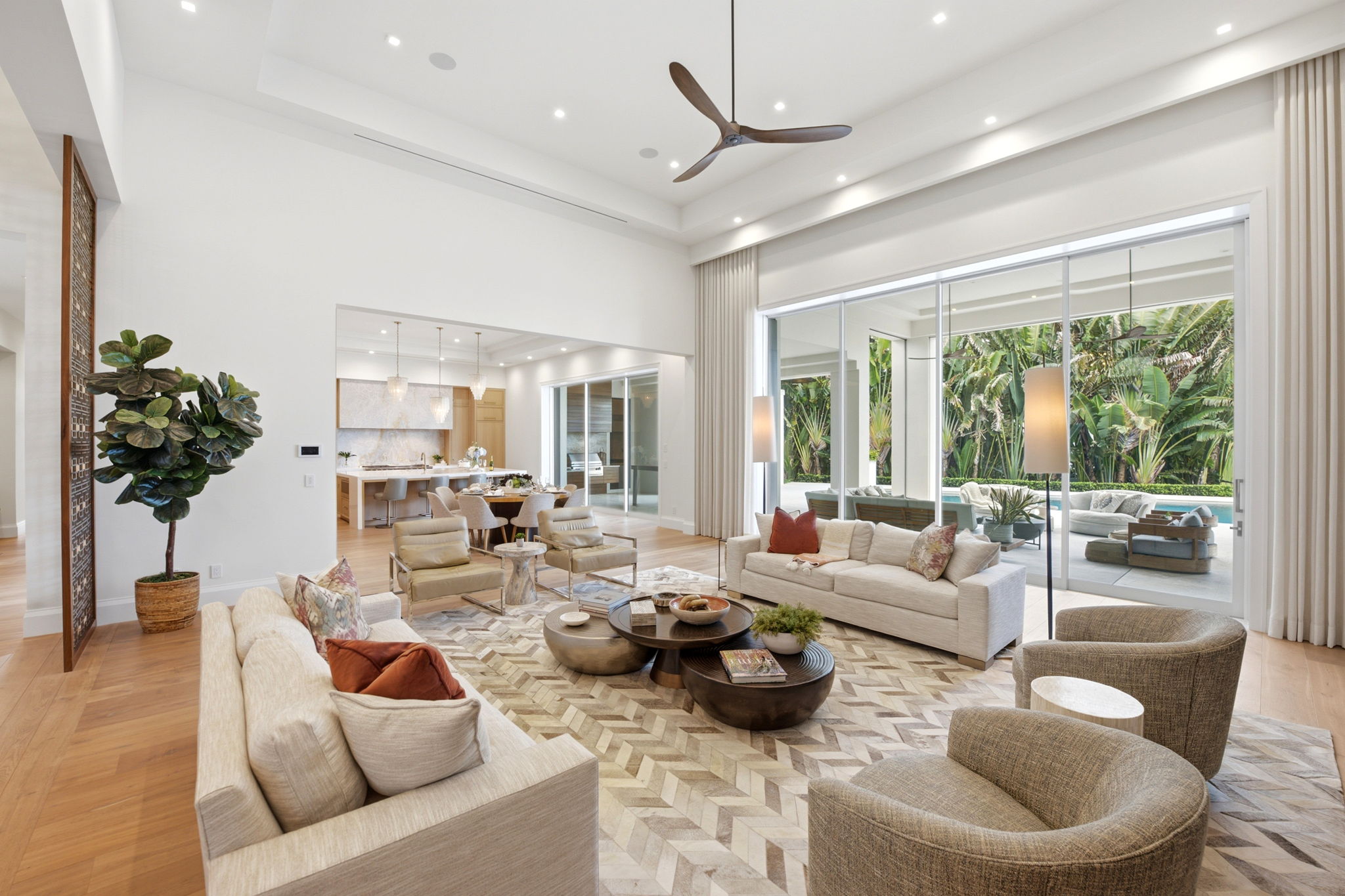 Bright, modern living room with beige sofas featuring colorful pillows, two beige armchairs, and two round coffee tables on a patterned rug. Large glass sliding doors open to a patio with outdoor seating and lush greenery. Ceiling fan with wooden blades hangs from the white ceiling with recessed lights. Adjacent dining area with a wooden table, beige chairs, and a kitchen with pendant lights visible in the background. A large potted plant stands near the entrance with wooden decorative paneling.