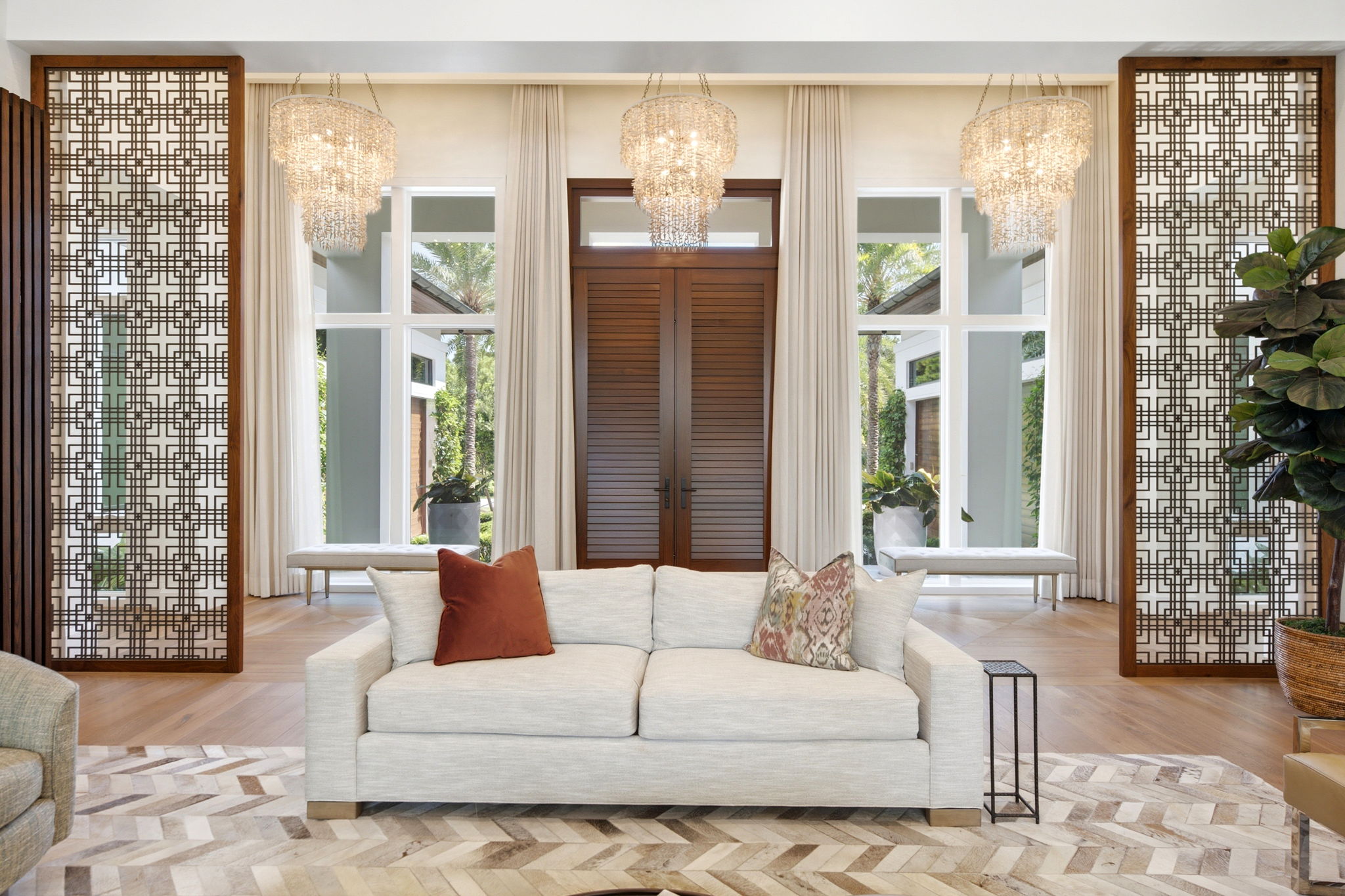 Bright modern living room with a beige sofa adorned with three decorative pillows, positioned on a patterned rug, in front of tall windows and wooden shutters, flanked by intricate wooden lattice panels and crystal chandeliers.