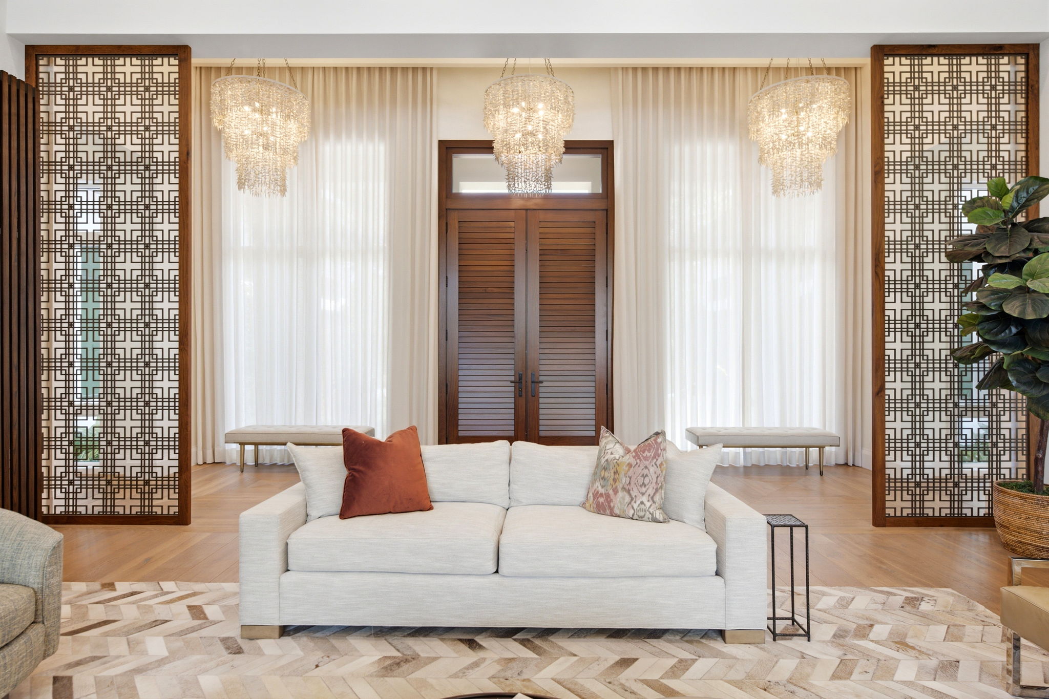 Modern living room with a light gray sofa featuring three decorative pillows, flanked by patterned wooden panels and two chandeliers hanging from the ceiling in front of tall white curtains and shuttered double doors.