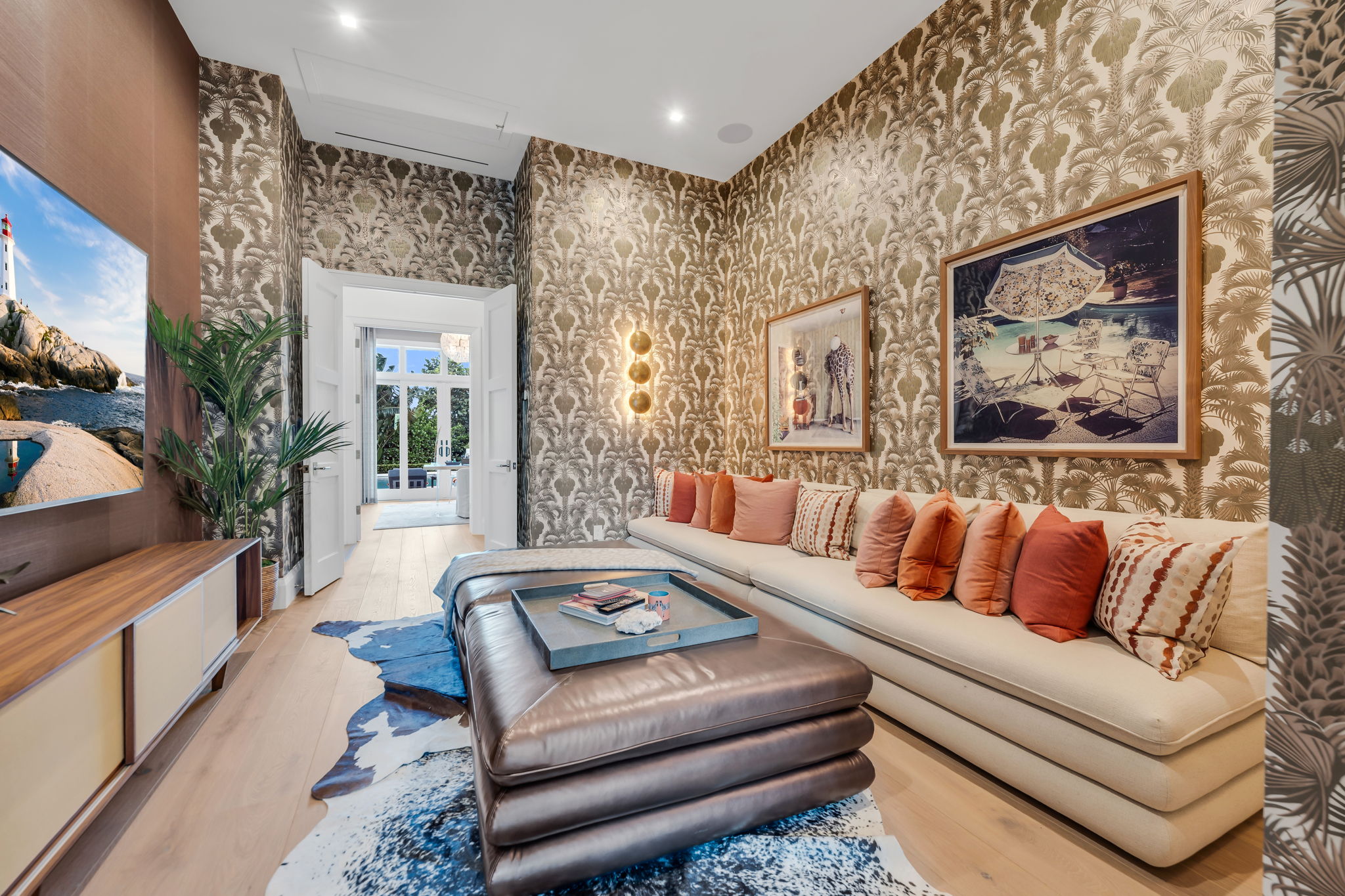Modern den with beige sofa adorned with assorted orange, peach, and patterned pillows, a layered brown leather ottoman topped with a tray and books, animal print rug on light wooden floor, tropical patterned wallpaper, two framed art pieces, gold wall sconces, and a TV displaying a lighthouse scene.