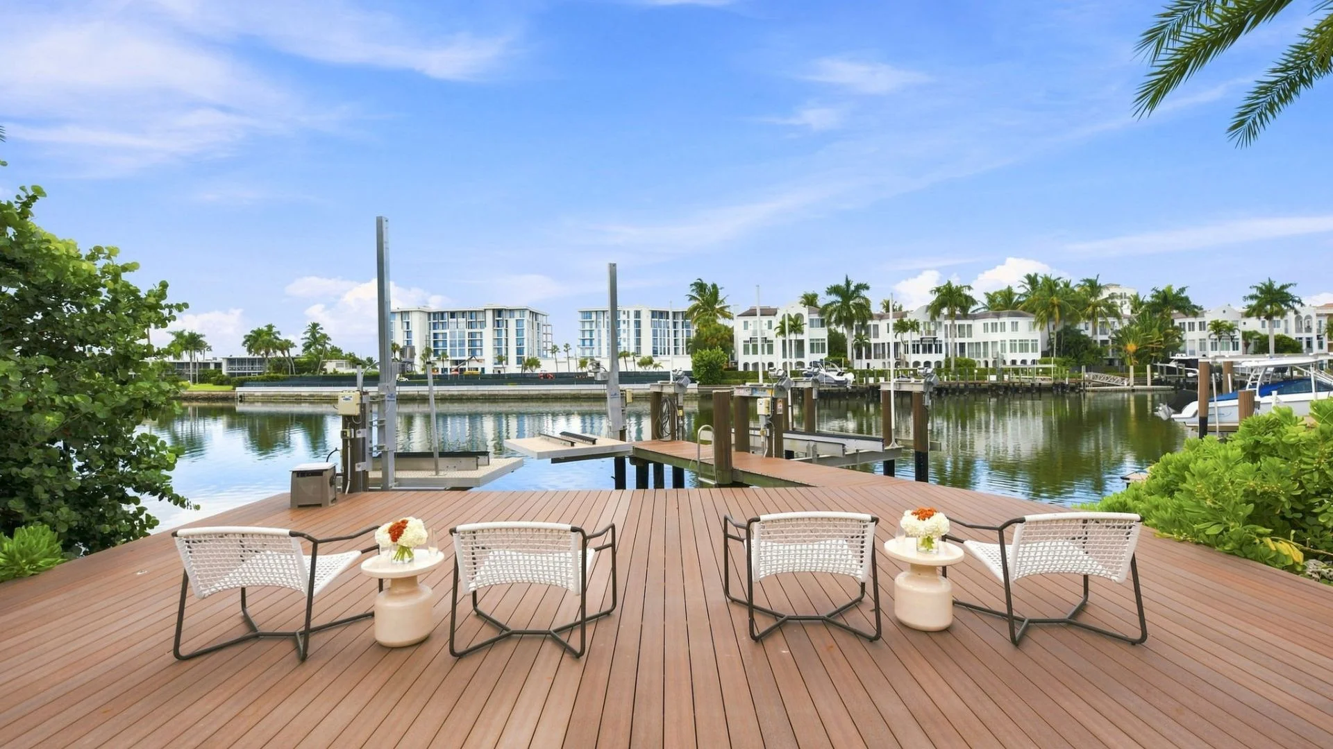 Wooden dock with four white woven lounge chairs and two small round tables with flower arrangements, overlooking a calm waterfront with boats and white buildings surrounded by palm trees under a partly cloudy blue sky.