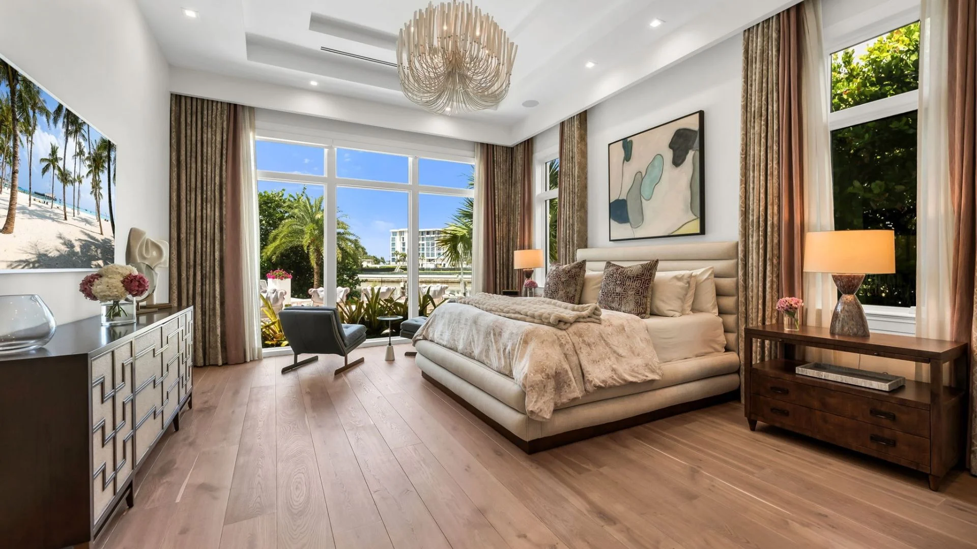 Modern bedroom with large bed featuring beige bedding and patterned pillows, two wooden nightstands with lamps, a stylish chandelier, large windows with a view of greenery and outdoor seating, hardwood floors, and a dresser with decorative items and a wall-mounted TV showing a beach scene.