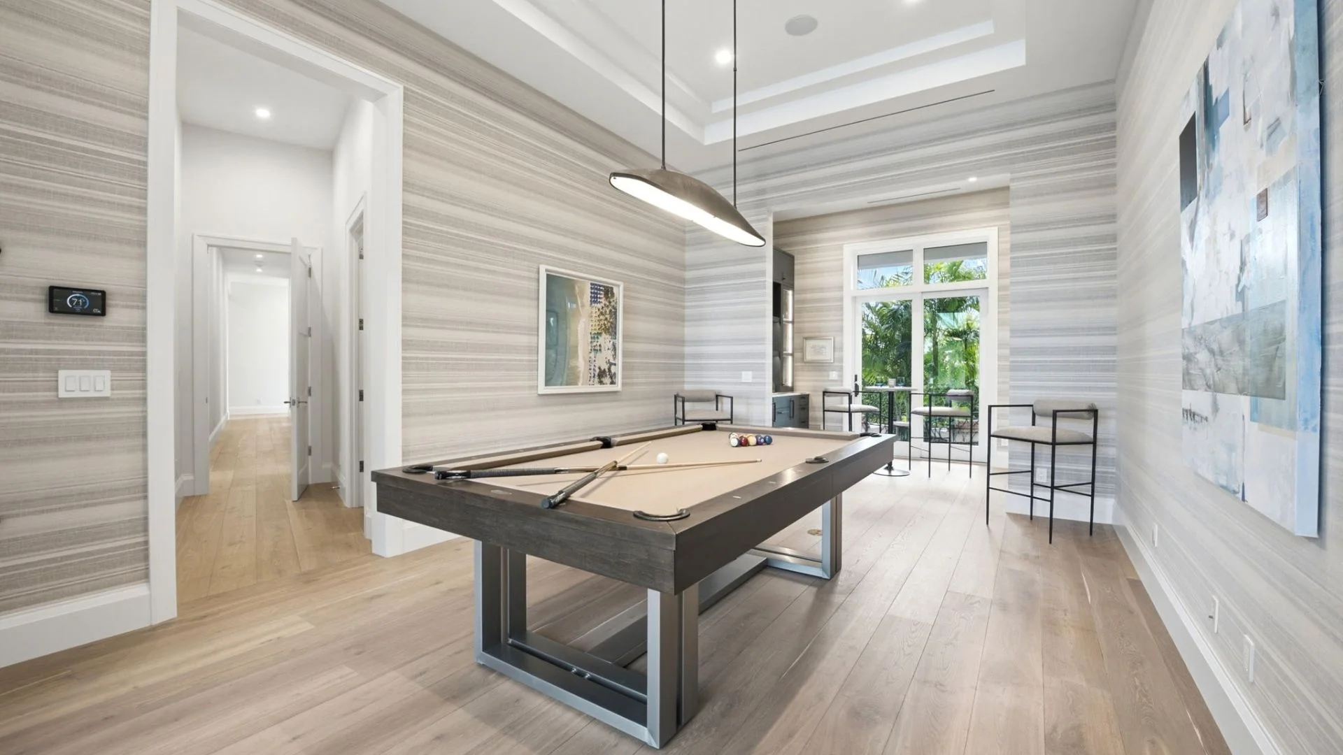 Modern game room with a beige pool table centered under a sleek pendant light, surrounded by light wood flooring, striped wallpaper, two tall chairs, artwork on the walls, and glass doors opening to a balcony with greenery outside.